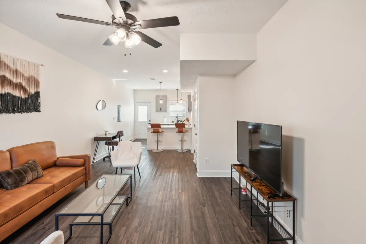 A spacious open-concept interior featuring a comfortable sofa, coffee table, TV, dining table, and kitchen island. The arrangement allows for easy movement and interaction, all bathed in natural light. The space combines living, dining, and kitchen areas, emphasizing both relaxation and social connection.