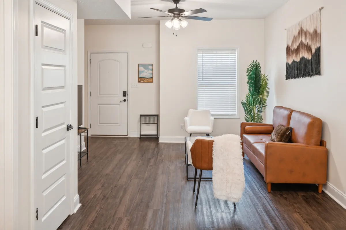 The image showcases a stylish, modern living room featuring a supple brown leather sofa, a chic white accent chair, lush green indoor plant, and contemporary wall art. The space is bathed in soft natural light, creating a warm, welcoming atmosphere perfect for relaxing or socializing. Decorative touches like framed pictures and thoughtfully arranged furnishings underscore a sense of comfort and sophistication.