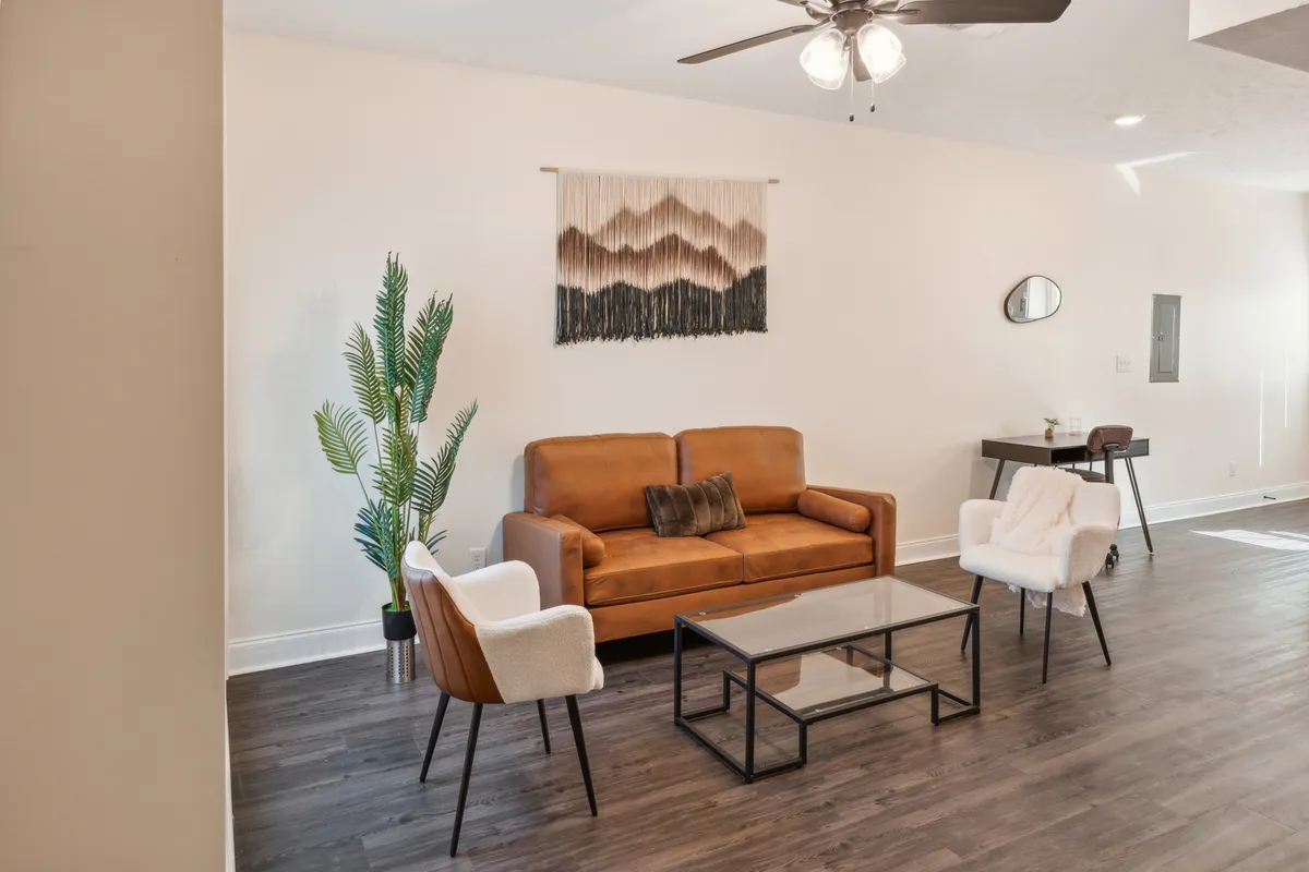 A cozy living room with a brown leather sofa, glass coffee table, textile wall hanging, potted plant, and white accent chairs. The space is designed for comfort and casual conversation, with inviting textures and a soothing ambiance.