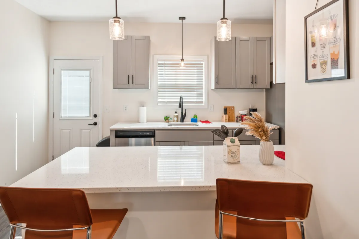 A contemporary kitchen space featuring a central island, stylish hanging light fixtures, a window with blinds that hint at outdoor light, modern cabinetry, and a decorative vase. The setting is open and inviting, ideal for meal prep and family gatherings.