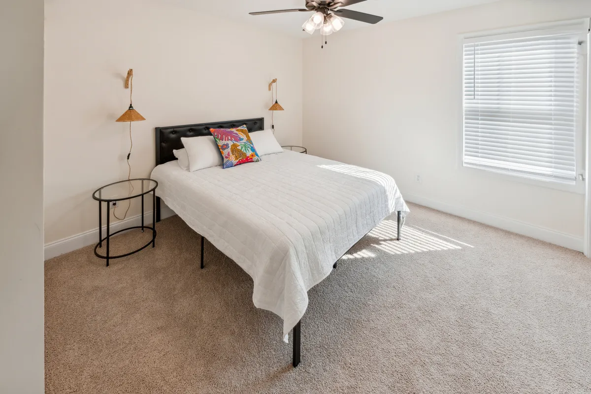 A cozy bedroom with a bed adorned with a colorful pillow, a ceiling fan overhead, and a nightstand with a lamp. Natural light streams in, creating an inviting, tranquil atmosphere that emphasizes relaxation after a busy day exploring Clarksville.