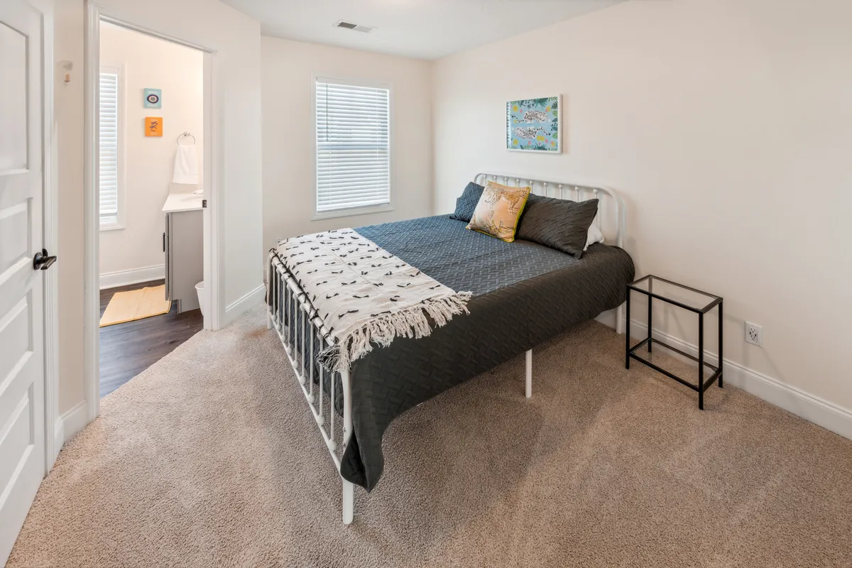 A cozy and inviting bedroom with a decorative throw on the bed, artwork on the wall, and a functional nightstand. The morning light creates a warm, welcoming mood, emphasizing comfort and personal space.